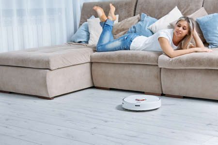 A pretty woman lies in jeans and a white T-shirt on the couch, she is happy because now a robot vacuum cleaner performs cleaning for herの写真素材