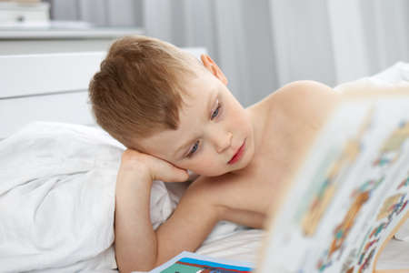 Little boy reading a book in a white bedの写真素材