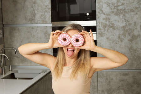 Portrait of a young woman smiling and covering her eyes with pink and white doughnuts in the kitchen