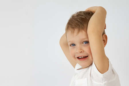 A happy child with blue eyes threw his hands behind his head on a white background. The boy smiles...