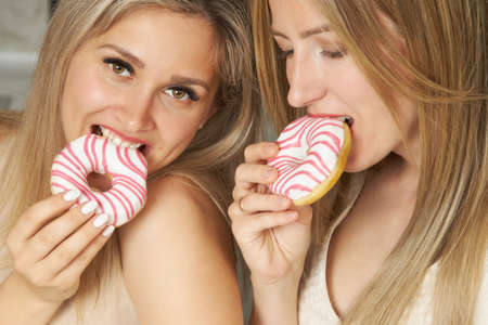 Two charming blonde women in evening dresses sexy bite off pink and white glazed donutsの写真素材