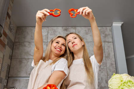Two cute blonde women have fun while cooking in the kitchen. The girls lifted the slices of red sweet pepper on their headsの写真素材