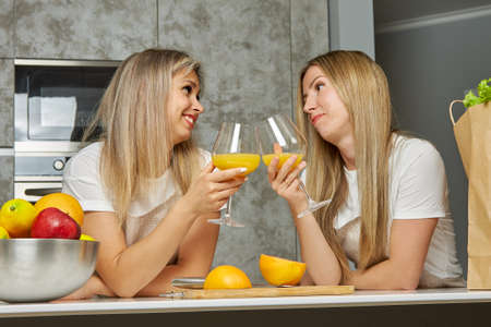 Two cute blonde women are drinking an orange alcoholic cocktail from glasses in the kitchen and...