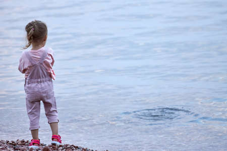Cute little blonde girl in a pink jumpsuit stands alone on the beach and looks into the distance.の写真素材