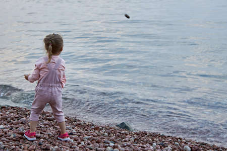 Cute little blonde girl in a pink jumpsuit stands alone on the seashore and throws pebbles into the waterの写真素材