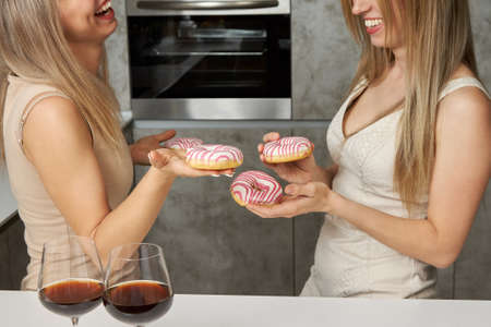 Delicious doughnuts in white and pink glaze on the hands of two beautiful blondes in evening dresses