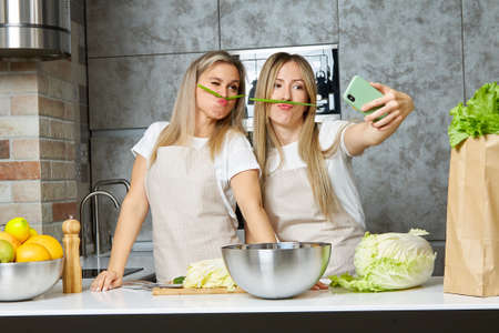 Two women cook food, make themselves a mustache from onions, have fun and take selfies in a modern...