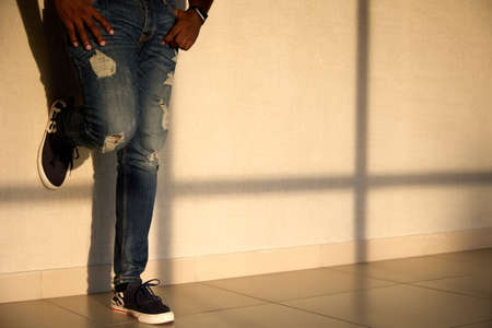 Cool black man in jeans and sneakers stands in the sunlight in front of the windowの写真素材