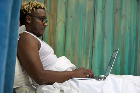 Black awake man in a white T-shirt and glasses is working on a modern laptop in bed. Always online.