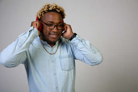 African-American man is smiling in a blue shirt and glasses, listening to red wireless headphones on...