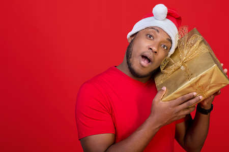 Excited black man with his mouth open and wearing a Santa Claus hat, shaking a packed gift box, is interested in what is inside, receives Christmas gifts, standing on a red backgroundの写真素材