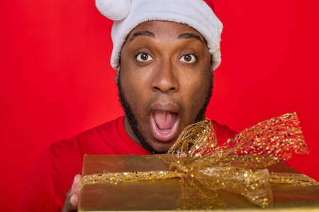 Close-up of a cheerful black guy with an open mouth in a Santa hat giving a Christmas gift in a golden package on a bright red backgroundの写真素材