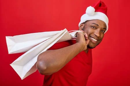 Close-up of a black man in a Santa hat who carries a white paper bag on his back like a bag. The concept of buying gifts at the Christmas sale on a bright red backgroundの写真素材