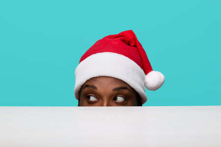 African-American man in a Santa Claus hat with expressive eyes looks carefully to the left looking out from under a white table on a turquoise backgroundの写真素材