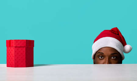 African-American man in a Santa Claus hat with expressive eyes looks attentively at the camera looking out from under a white table on which there is a beautifully packaged giftの写真素材