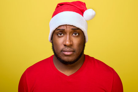 Portrait of an attractive black guy in a Christmas hat and a red T-shirt confidently looking directly into the camera isolated on a bright yellow backgroundの写真素材