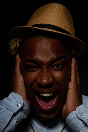 African-American man in a straw hat and a blue shirt, crazy and scared, holds his head in his hands with his mouth open from shock on a black backgroundの写真素材