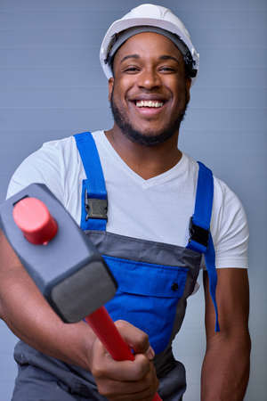 An African-American man in a white helmet and work clothes smiles with a toothy smile, holding a sledgehammer in his hands on a gray background. Vertical portrait of a happy worker in overallsの写真素材