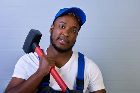 Black man in a cap and work clothes is smiling, holding a hammer on his shoulder. Portrait of an African-American worker in overalls with a copy space.の写真素材
