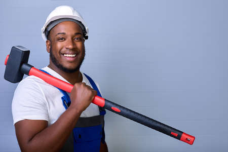 Professional construction worker in a protective helmet and work clothes looks at the camera, smiles and holds a sledgehammer on his shoulder. Portrait of a black worker in overallsの写真素材