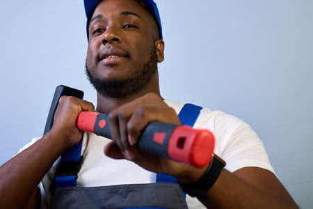 Black man in a cap and work clothes holds a hammer in his hands. Close-up of a worker in overalls with a sledgehammer in his hands on a gray backgroundの写真素材