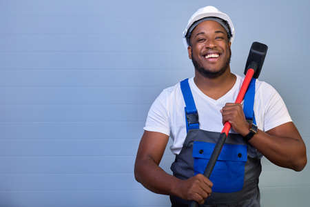 African-American man in a white helmet and work clothes smiles with a toothy smile, holding a sledgehammer in his hands on a gray background. Portrait of a happy worker in overalls with a copy spaceの写真素材