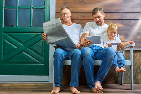 Grandfather with a newspaper, a son with a tablet, a grandson with a smartphone are sitting on a...