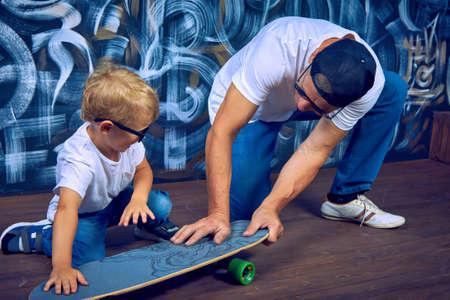Cool grandfather in cap and sunglasses is having fun together with his grandson riding skateboard. Modern elderly man actively spends time with his grandson the child has lot of fun playing with himの写真素材