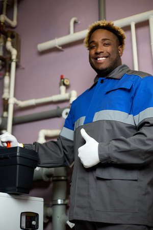 Smiling African-American worker in overalls stands in the boiler room and holds a suitcase with special tools. Black man shows a thumbs up gesture indicating the high quality of the work carried outの写真素材