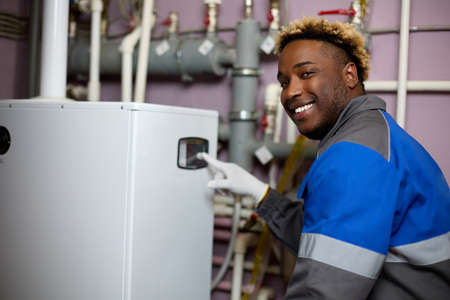Smiling African-American worker in overalls sets up a gas boiler, adjusts a hot water boiler. Black specialist presses his finger on the display of a floor boilerの写真素材