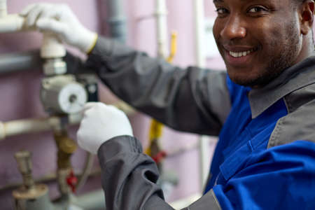 Happy professional black worker in overalls adjusts the sensors in the boiler room using a...