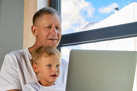 Happy grandfather and grandson are watching interesting content on a laptop using the Internet. Modern grandpa spends time with his grandchild using modern technologyの写真素材