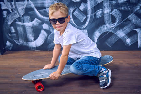 Pretty boy in sunglasses is sitting on a large skateboard. Little boy riding on a boardの写真素材