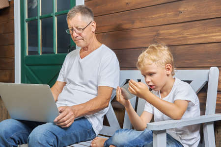 Grandfather with a laptop and a grandson with a smartphone are sitting on bench near a wooden house. Concept of using modern gadgets by different generationsの写真素材
