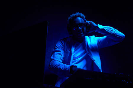 African-American DJ with headphones is sitting in a dark studio, a blue glow falls on him. DJ in a...