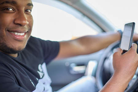 Smiling black man or a driver driving a car and using a smartphone. Concept of transport, vehicles...