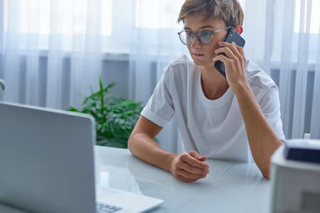 Handsome boy in a white shirt and glasses is talking on the phone and looking at a laptop. A...