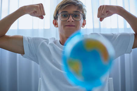 Cheerful teenager in glasses and a white T-shirt is fooling around and smiling at camera. Globe is...