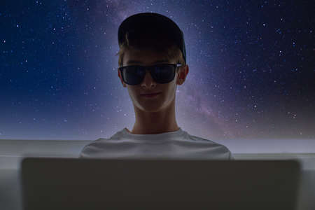 Teenager in dark glasses and a cap is sitting at a laptop against the background of stars. Space flights in the future. A teenager is studying space. A mysterious guy. Study of astronomy.の写真素材