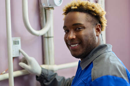 Black worker in overalls, smiling, adjusts a sensor hanging on the wall in the boiler room. African male adjuster changes the parameters of the equipmentの写真素材