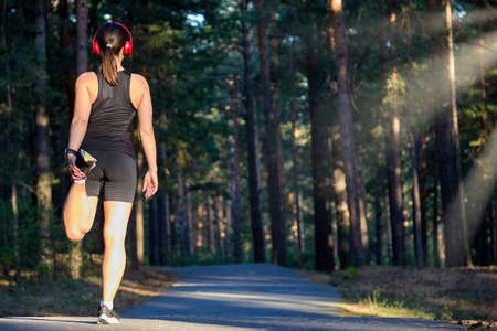 Athlete-runner in wireless headphones, stretching her legs and feet and preparing to run on an...