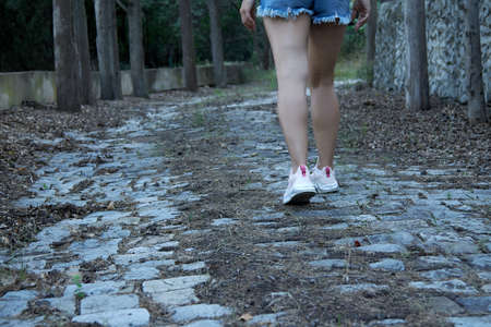 Traveler dressed in pink sneakers and blue denim shorts walks along a paved path. Beautiful womens...