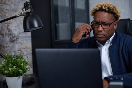 Serious African-American businessman with glasses is talking on the phone at the workplace, using a laptop, looking at the screen. Close-up of an employee consulting a client, solving a problemの写真素材