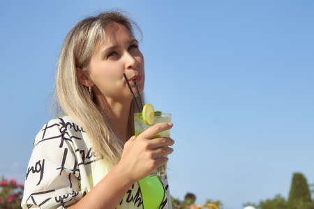 Beautiful young woman drinks a lime drink from a straw in hot weather. A refreshing drink on a clear day helps to cool down.の写真素材