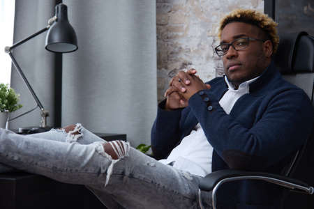 Serious confident African businessman in ripped jeans sits with his legs folded on the table and looks at the camera. Concept of a free lifestyle of an entrepreneur or freelancer.の写真素材