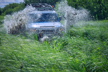 Off-road car crashed into a puddle and raised mud splashes. A four-wheel drive sports car is racing among the green grass. Off-road 4x4 SUV in blue color. The best off-road vehiclesの写真素材