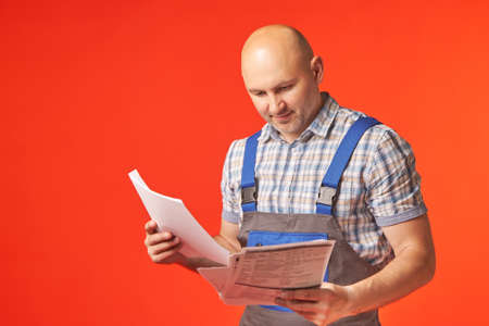 Bald man in work clothes holds in his hands and examines documents, a photo on a red background. The builder looks at the drawing to perform the work.の写真素材