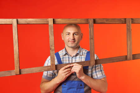 Bald man in work clothes holds a wooden ladder horizontally and looks through a frame, a photo on a red background. Rest at an employee with a ladder.の写真素材
