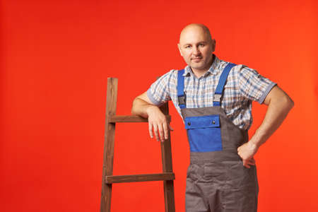 Bald man in work clothes is standing with his hand on the stairs, a photo on a red background. An employee is resting, holding onto a wooden ladder.の写真素材