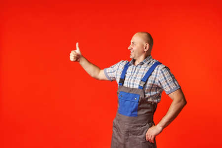 Smiling bald man in work clothes on a red background shows an ok gesture to the side, raising his hand. The chief evaluates the work of the builders.の写真素材